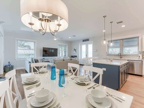 A modern kitchen with a central island, dining area with white table and chairs, and a living area with a flat-screen TV.