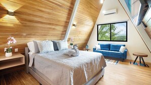 A modern bedroom with a sloped ceiling, a large bed with a swan decoration, a blue sofa, and a window with a view of greenery. - Resort Villa 5 Min to the Beach w Pool + Kitchen (Potrero)