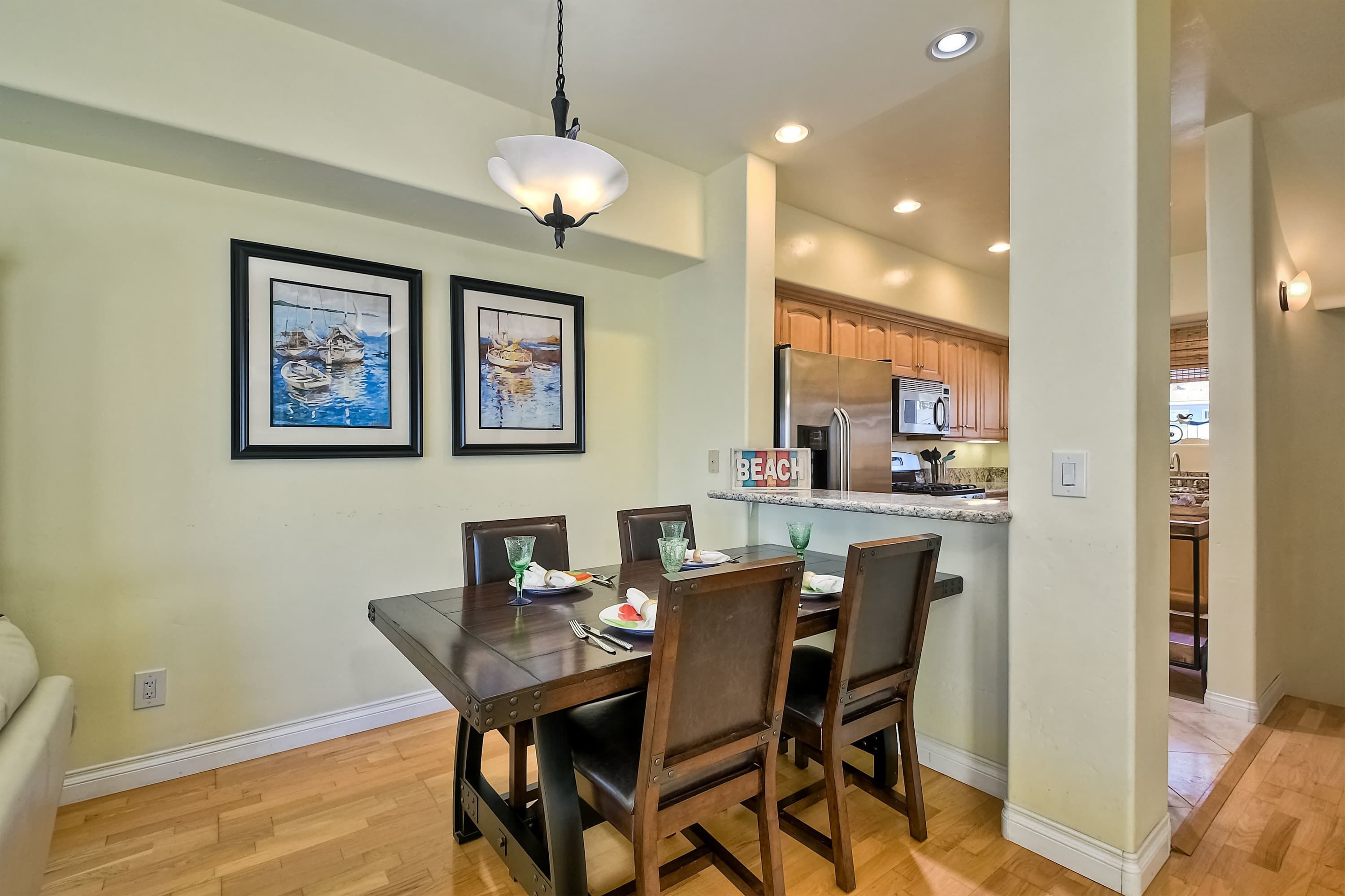 A dining area with a wooden table and chairs, framed artwork on the wall, and a kitchen in the background.