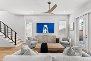 Un salon moderne avec une table basse en bois, deux fauteuils gris, un grand téléviseur à écran plat et un ventilateur de plafond.