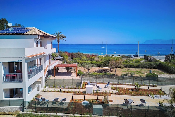 A modern building with a rooftop solar panel, a pool area with lounge chairs, and a view of the sea.