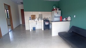 Cocina compacta con refrigerador, estufa y lavamanos. Un cesto azul está en el suelo y un sofá negro en la sala.