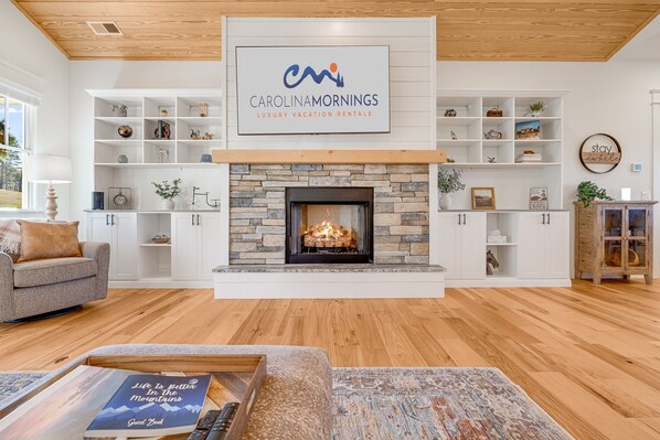 Una sala de estar moderna con chimenea de piedra, estanterías blancas con diversos objetos decorativos, y un cartel de Carolinamornings Luxury Vacation Rentals.