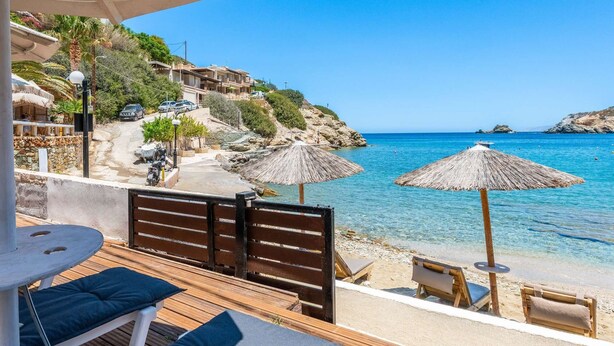 Een houten terras met lounge fauteuils en parasols met uitzicht op een heldere blauwe zee, met gebouwen en groen op de achtergrond.