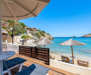 Een houten terras met lounge fauteuils en parasols met uitzicht op een heldere blauwe zee, met gebouwen en groen op de achtergrond.