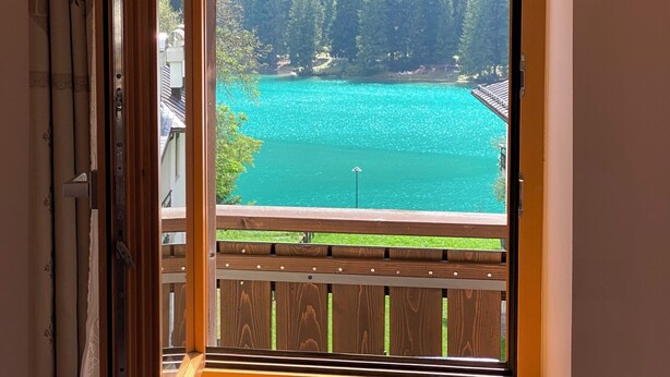 Een kamer met een groot raam, uitzicht op een turquoise meer omringd door dicht bos.