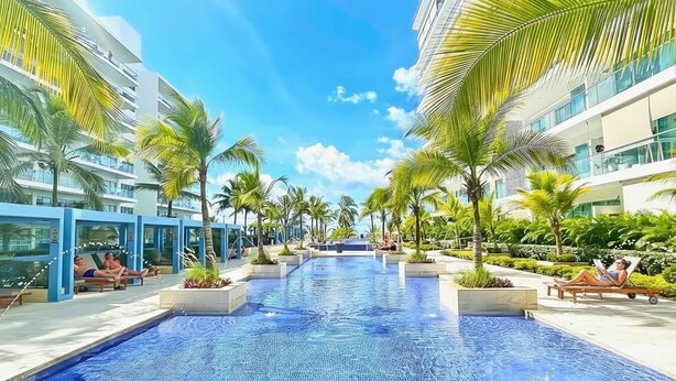 A pool area with lounge chairs and palm trees, surrounded by modern buildings.