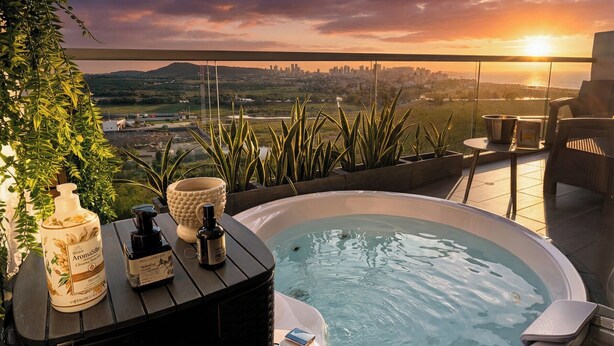 A hot tub with a view of a city skyline at sunset, surrounded by plants and a small table with toiletries.