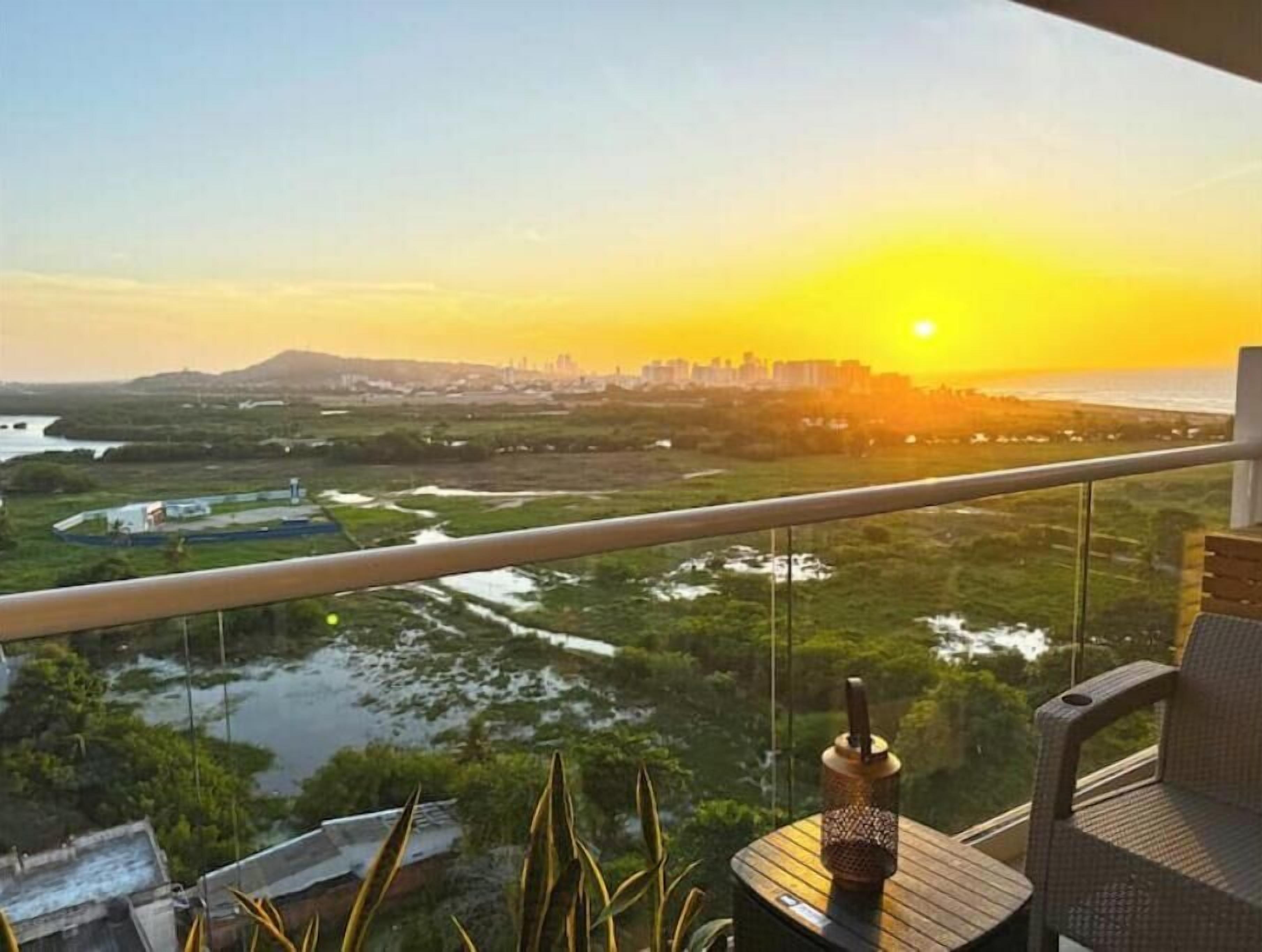 A balcony with a view of a city skyline at sunset, a body of water, and a table with a decorative item.