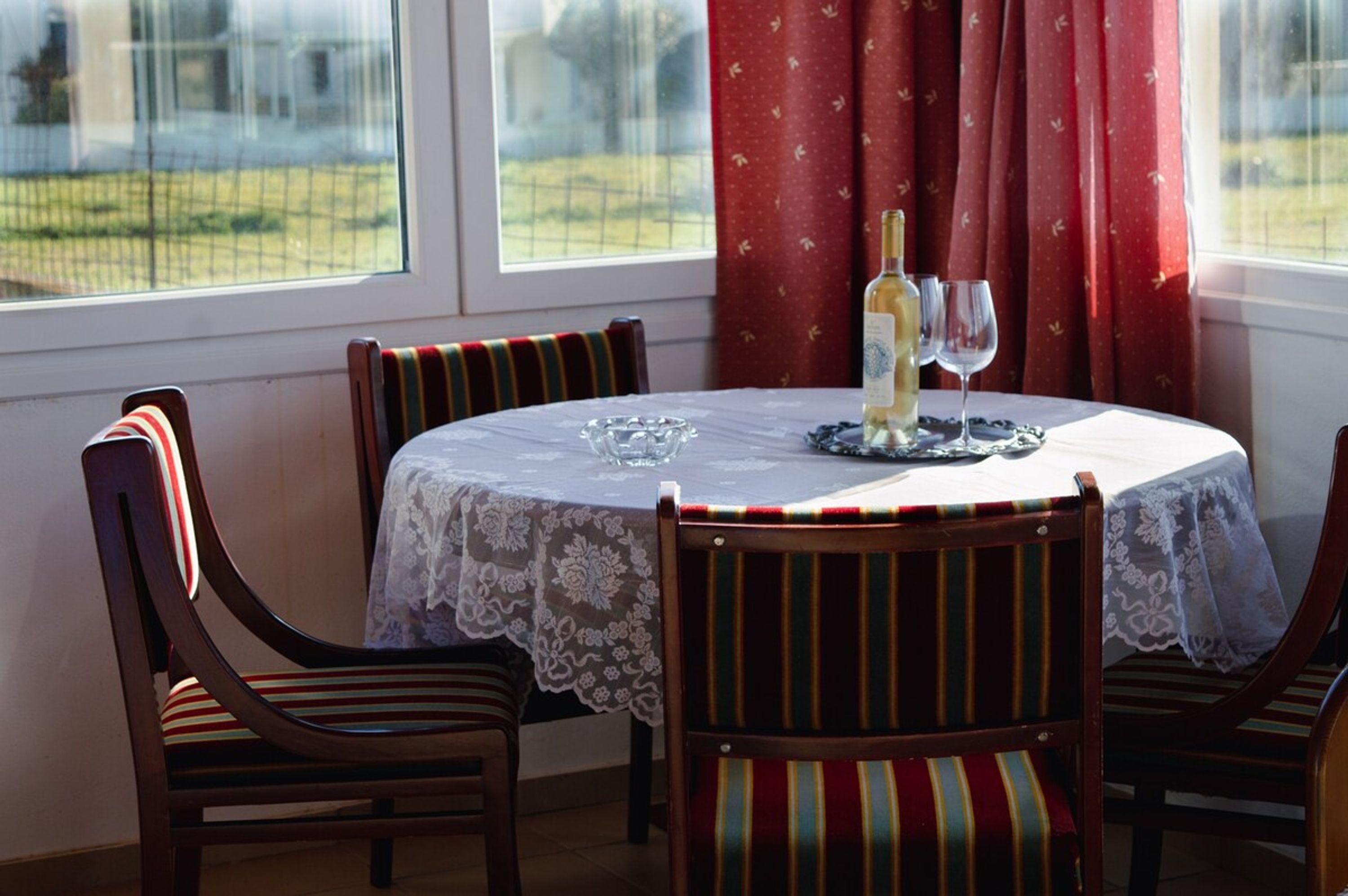 A dining table with a lace tablecloth, four wooden chairs with striped cushions, a bottle of wine, and two wine glasses on the table.