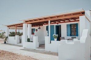 Un edificio blanco con una pérgola de madera, contraventanas azules y plantas en macetas.