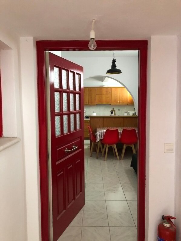 A red-framed door leading into a kitchen with wooden cabinets, a dining table with red chairs, and a tiled floor.