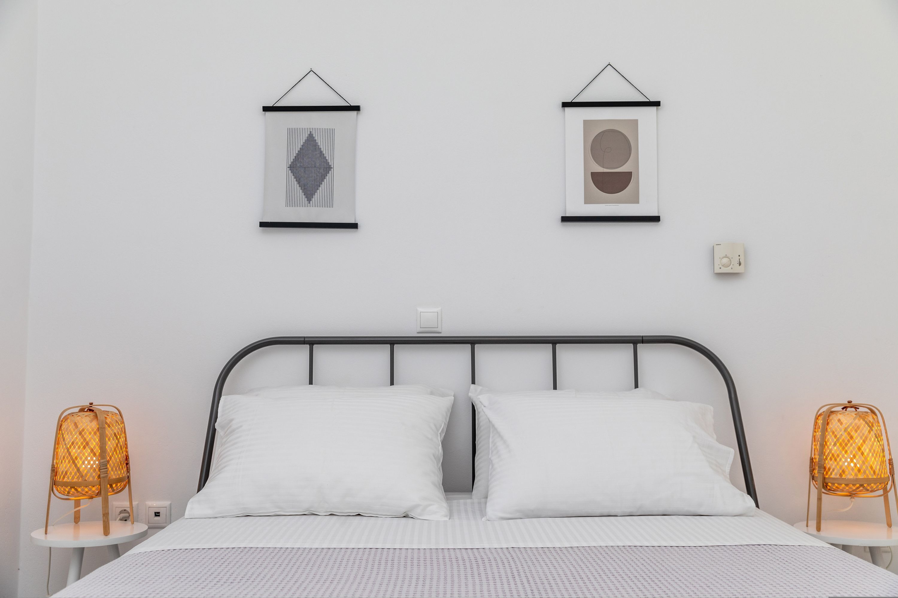 Een netjes opgemaakt bed met witte lakens, twee ingelijste kunstwerken aan de muur en twee rieten lampen aan weerszijden.