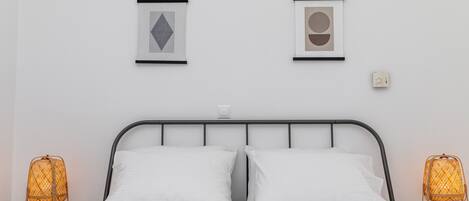 A neatly made bed with white linens, two framed artworks on the wall, and two wicker lamps on either side.