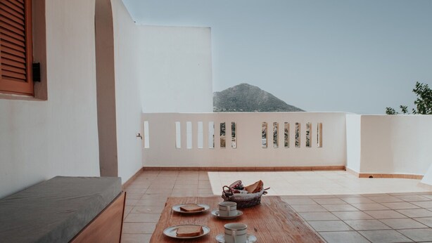 A balcony with a wooden table set for breakfast, a sofa, and a view of a mountain. - Tsikalario Holiday House (Naxos)