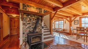 A cozy room with a stone fireplace, wooden beams, a staircase, and a dining area.