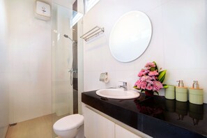 A modern bathroom with a glass shower enclosure, a white toilet, a black countertop, a round mirror, and a vase with pink flowers. - Villa Kyra Canggu (Canggu)