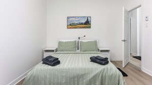 Un dormitorio con una cama, dos almohadas, una mesita de noche, un cuadro en la pared y una puerta.