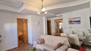A living room with a beige sofa, a TV mounted on the wall, a wooden door, and a dining area with a table and chairs. - weforyou Solymar 2 bedrooms (Benalmádena)