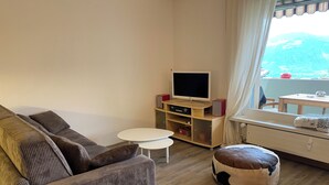 Ein Wohnzimmer mit einer Couch, einem kleinen weißen Tisch, einer TV-Konsole mit Elektronikgeräten, einem Fenster mit Blick und einem Holzboden.