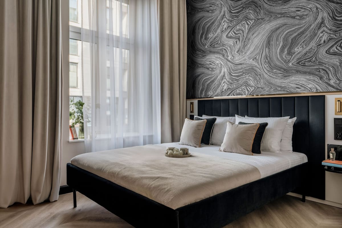 Une chambre d’hôtel moderne avec un grand lit, un mur décoré d’un motif imitant le marbre et une vue sur la ville à travers de fines rideaux.