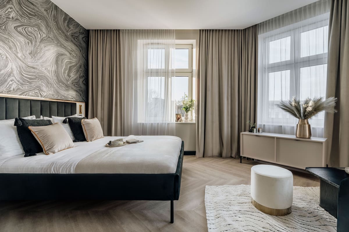 Une chambre moderne avec un grand lit, une table de chevet, une fenêtre avec des rideaux et un mur orné d’un motif évoquant le marbre.