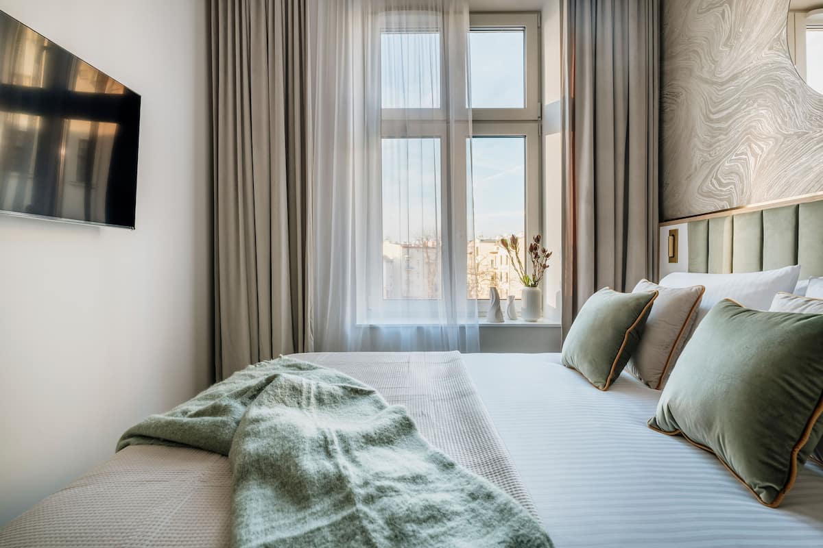 Une chambre d’hôtel avec un grand lit, un téléviseur à écran plat, une fenêtre avec des rideaux légers et une vue sur les bâtiments environnants.