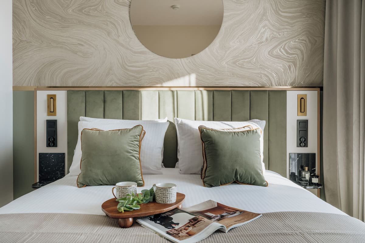 Un lit bien fait, avec des oreillers verts et blancs, un plateau avec des tasses et un magazine, et une table de chevet en marbre.
