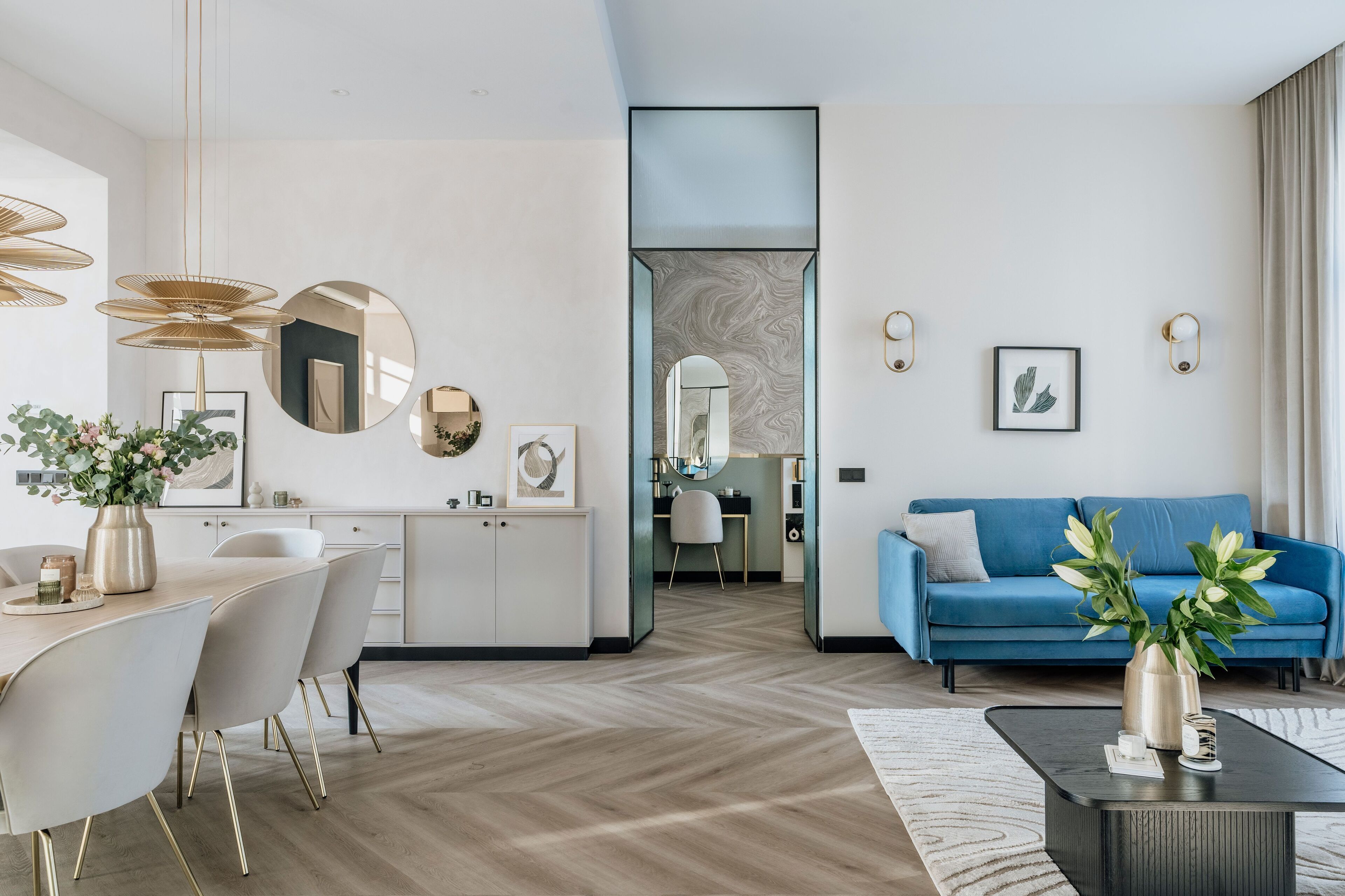 A modern living room with a blue sofa, a dining area with a white table and chairs, and a kitchen area with white cabinets and a large mirror.