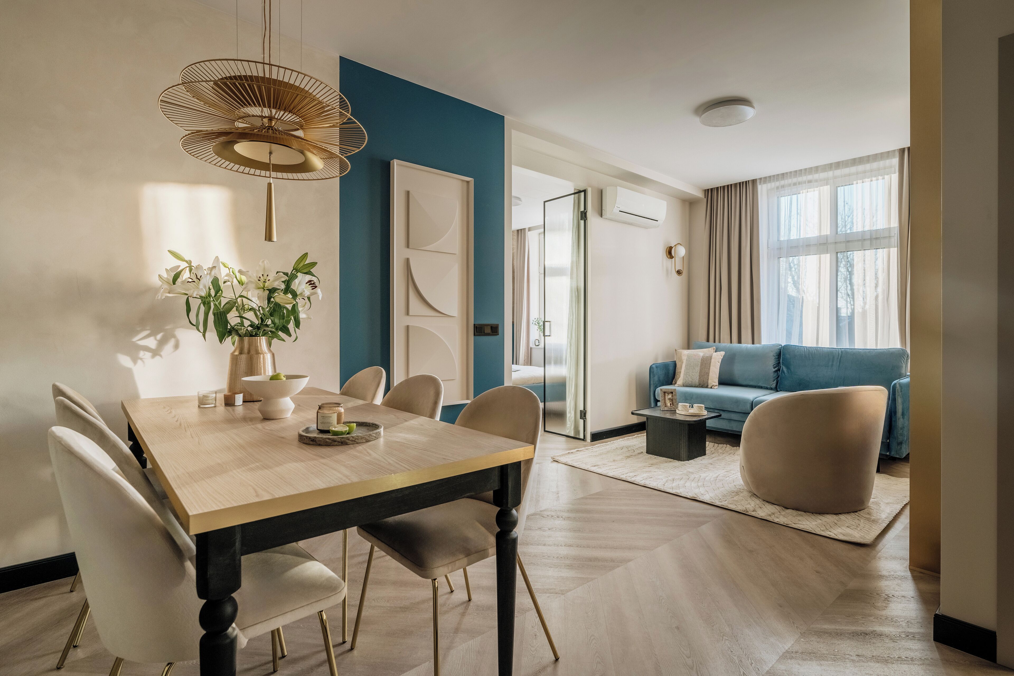 Una sala de estar moderna con comedor, que incluye una mesa de comedor de madera, sillas color beige, un sofá azul y un amplio ventanal con cortinas.