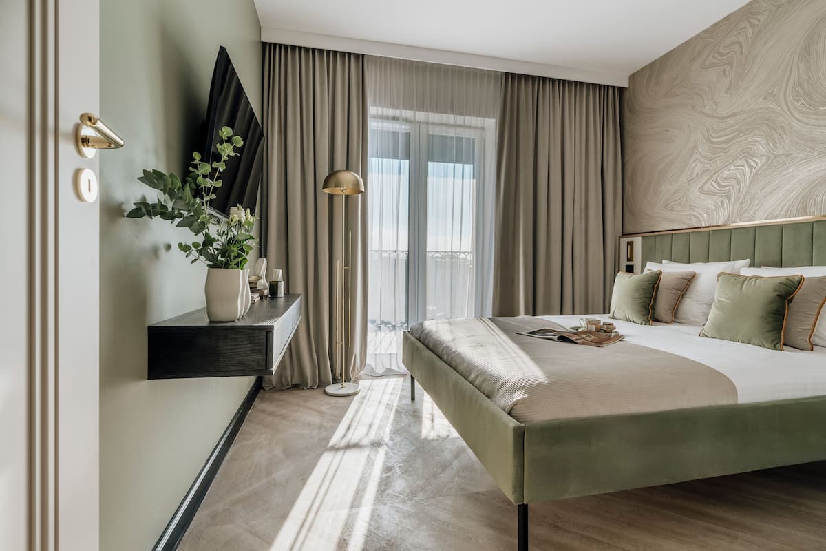 Une chambre d’hôtel moderne avec un grand lit, une table de chevet ornée d’un vase de fleurs, une lampe sur pied et une vue sur la ville par la fenêtre.