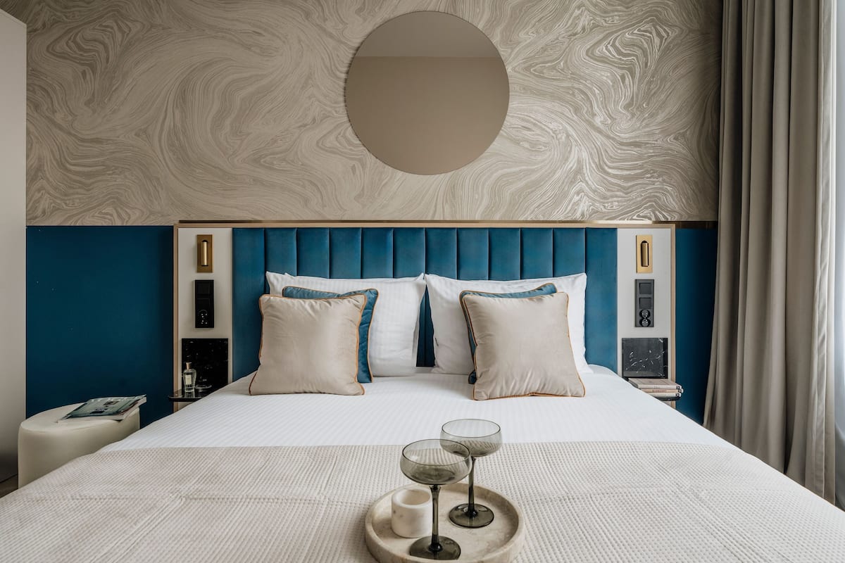 Une chambre d’hôtel moderne avec un plafond à motifs de marbre, une tête de lit bleue et un miroir rond au-dessus du lit.