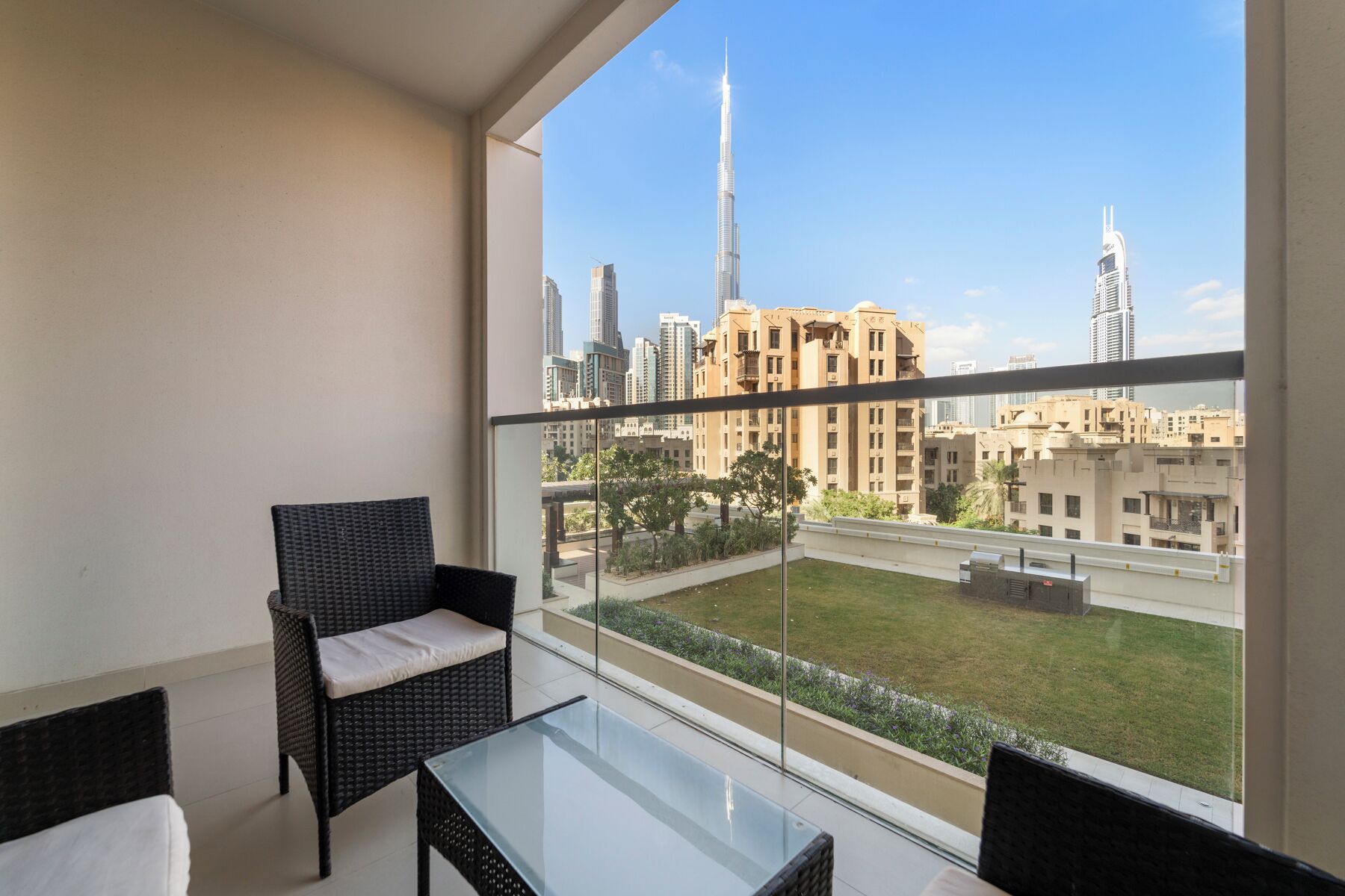 A balcony with wicker chairs and a glass table, overlooking a cityscape with modern buildings and a green lawn.