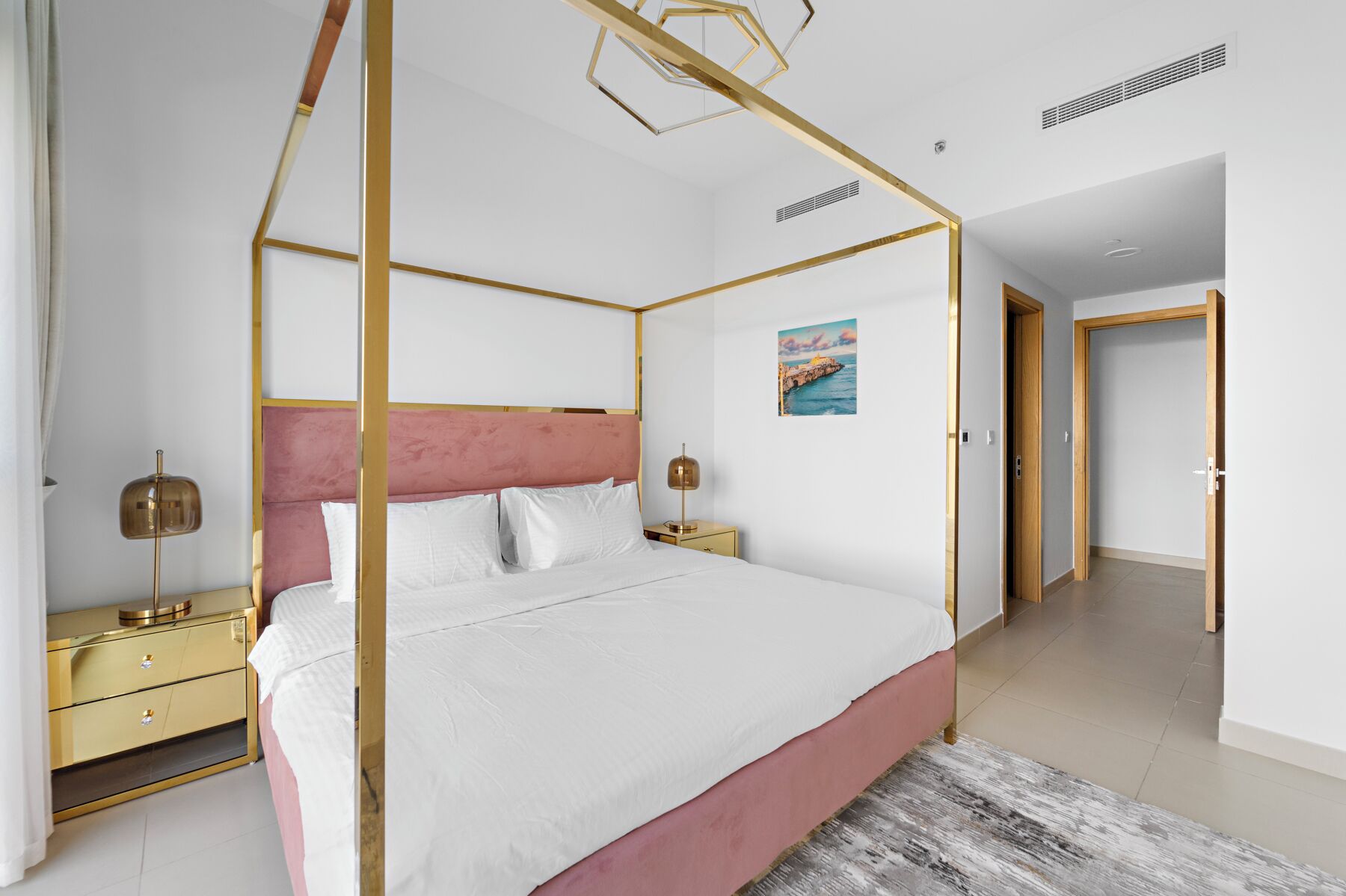 A four-poster bed with a pink headboard, white bedding, and gold accents. There is a nightstand with a lamp and a painting on the wall.