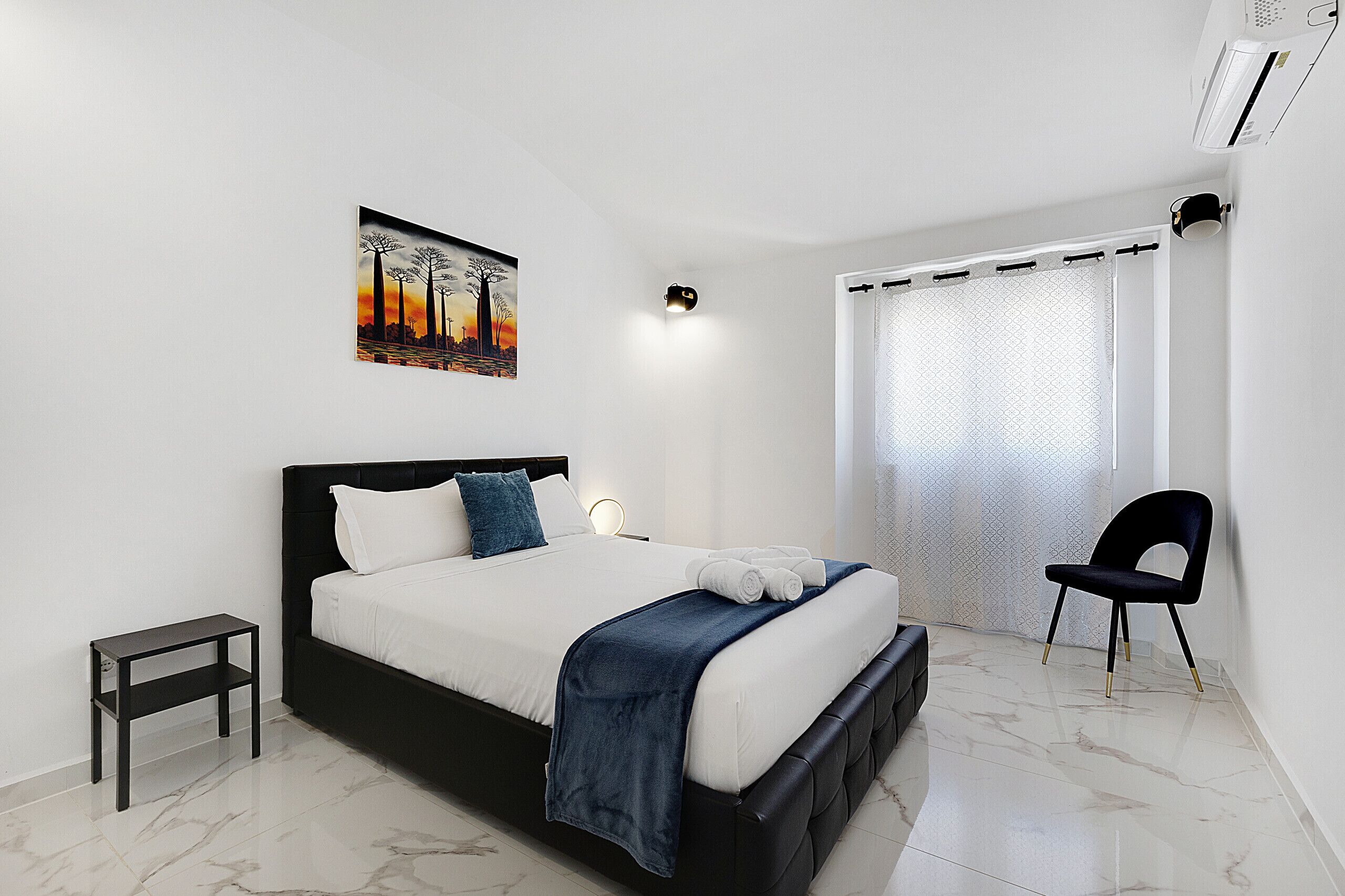 A modern bedroom with a bed, a small side table, a chair, a painting on the wall, and a window with a curtain.
