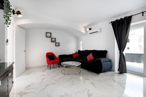 Una sala de estar con un sofá negro, una silla roja, una mesa de centro de vidrio y una pared blanca con un marco de fotos.