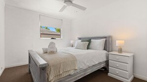 Un dormitorio con una cama, una mesita de noche, una lámpara, un ventilador de techo y una ventana con vista a un edificio.