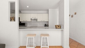 Cocina moderna con armarios blancos, microondas y cafetera. Hay dos taburetes en la barra.