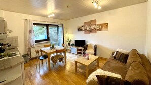 Una sala de estar moderna con piso de madera, un sofá marrón, una mesa de comedor con sillas, un televisor y una ventana con cortinas.