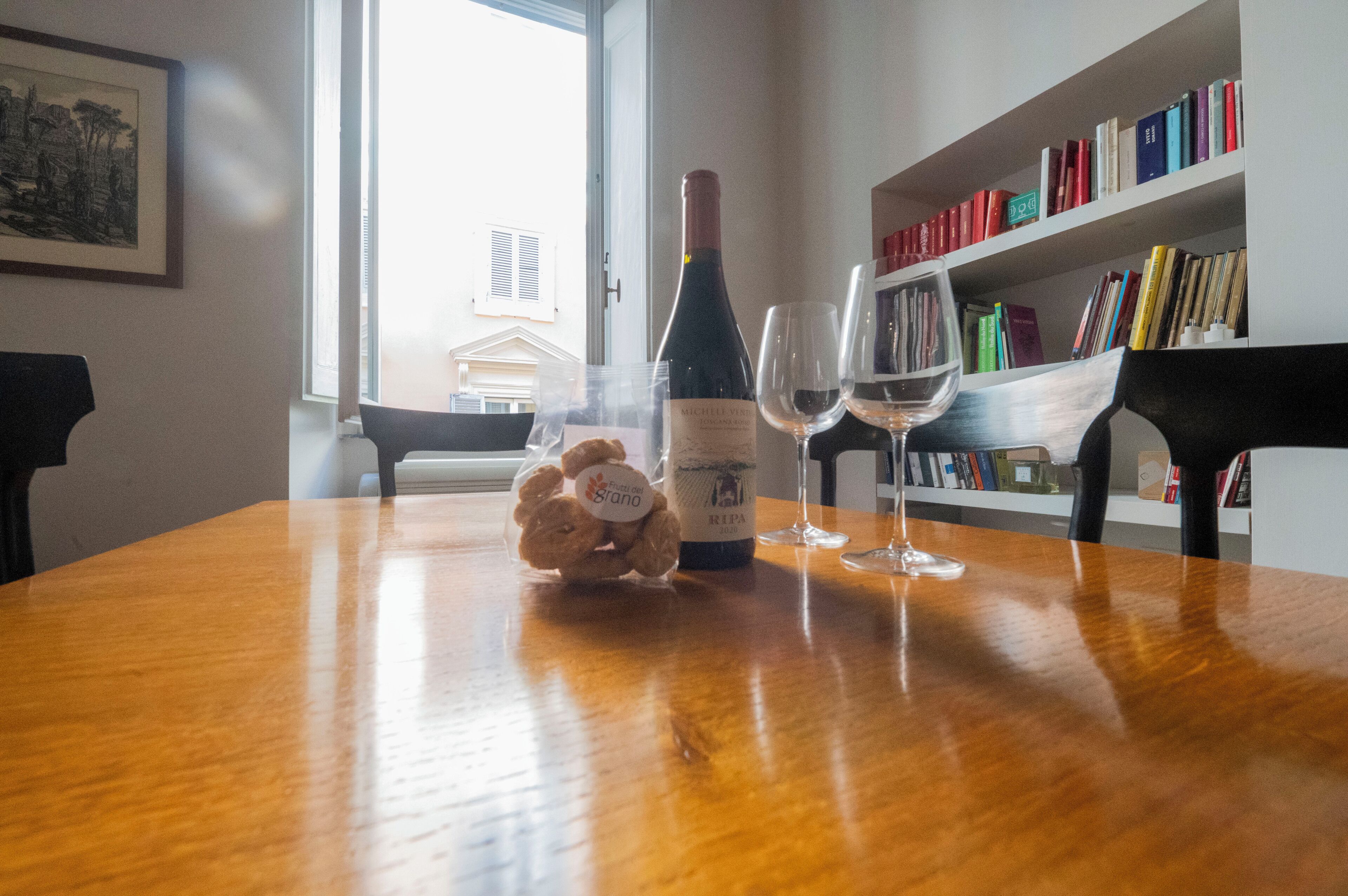 A wooden dining table with a bottle of wine, two wine glasses, and a bag of snacks.