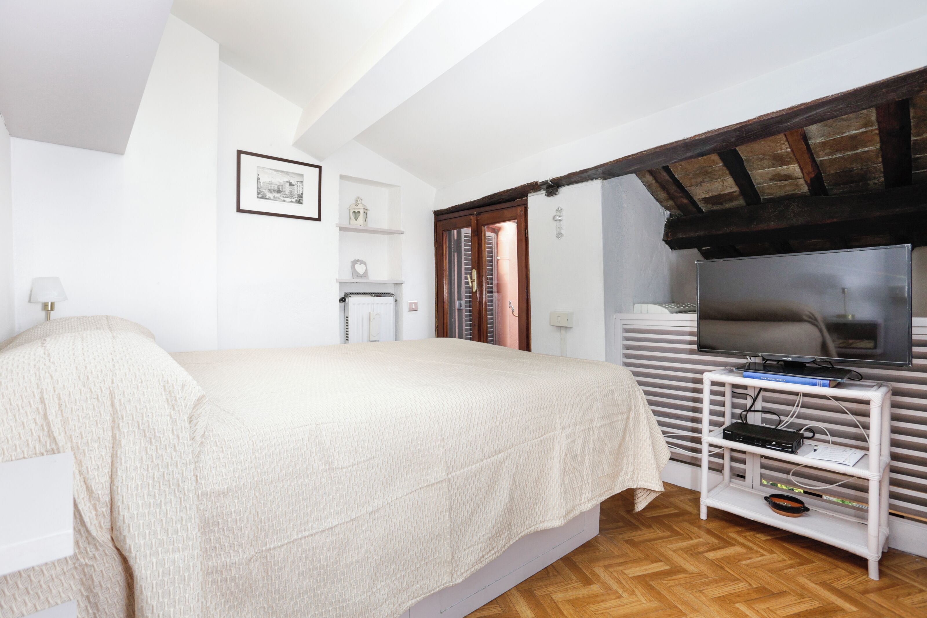 Une chambre à coucher avec un lit, un téléviseur posé sur un meuble, un plafond en bois et une fenêtre avec des stores.