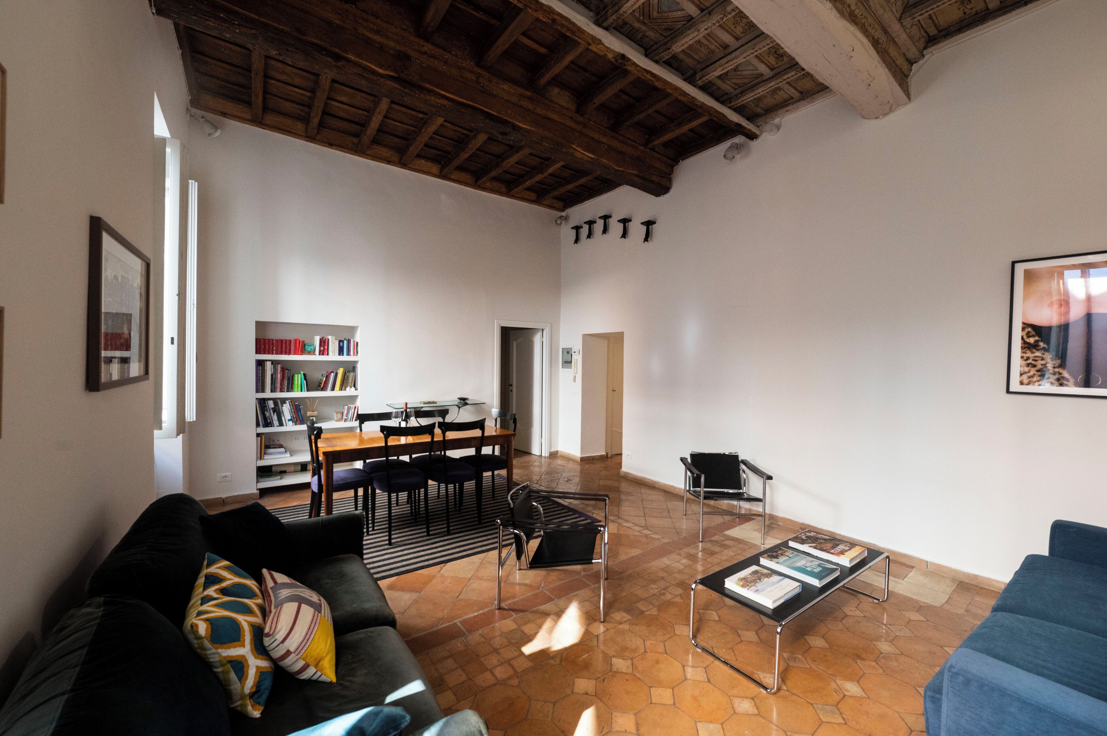 Een ruime woonkamer met een hoge plafond, een eethoek met een houten tafel en stoelen, en een boekenplank vol boeken.