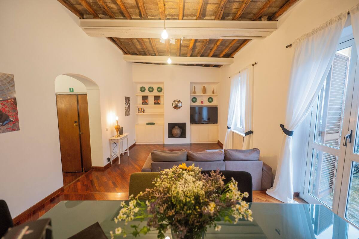 Un salon chaleureux avec un plafond en bois, un canapé, un téléviseur et un coin repas comprenant une table en verre et des fleurs.