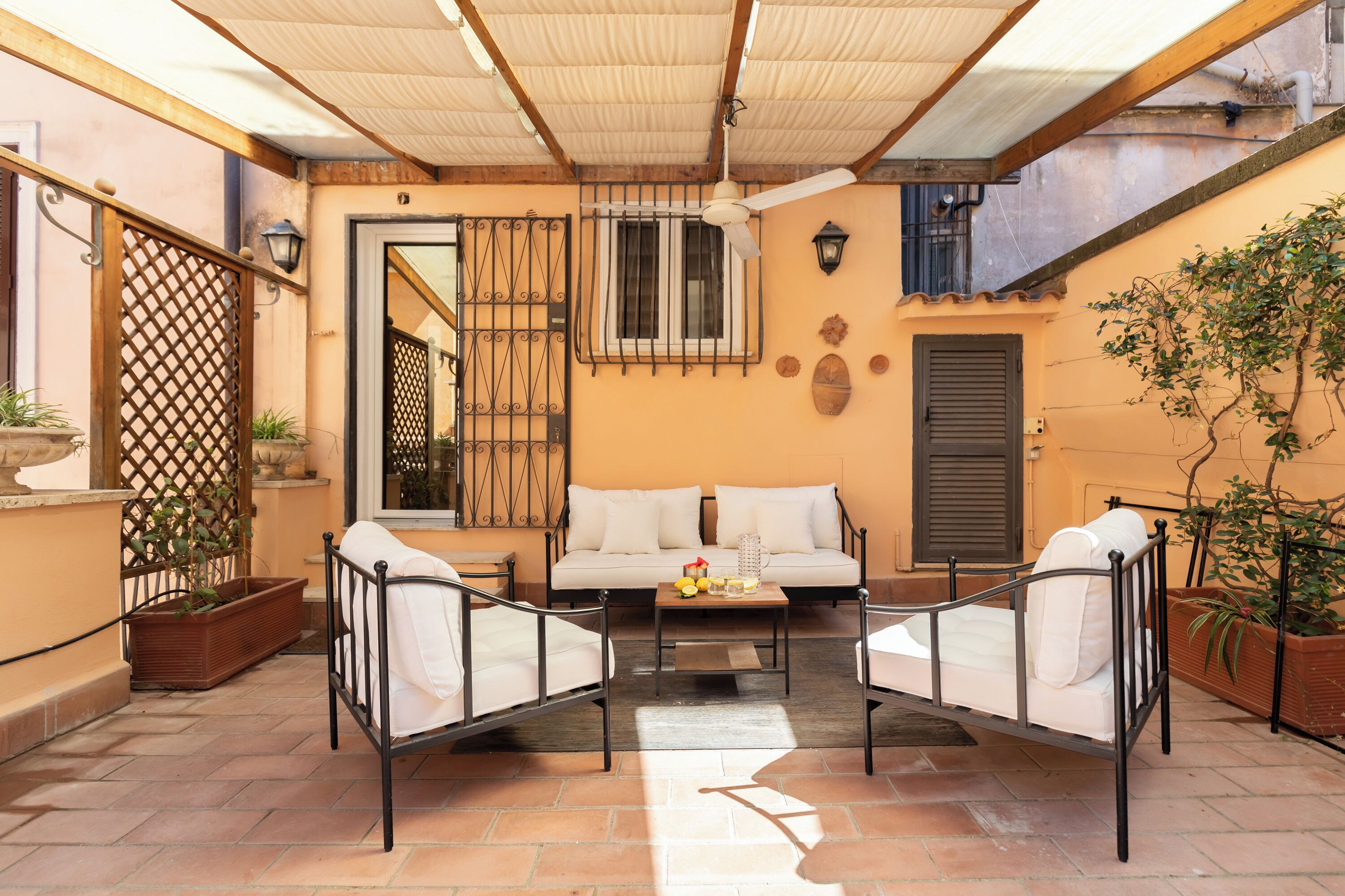 A cozy outdoor seating area with a sofa and two chairs, a small table with fruits, and a ceiling fan.