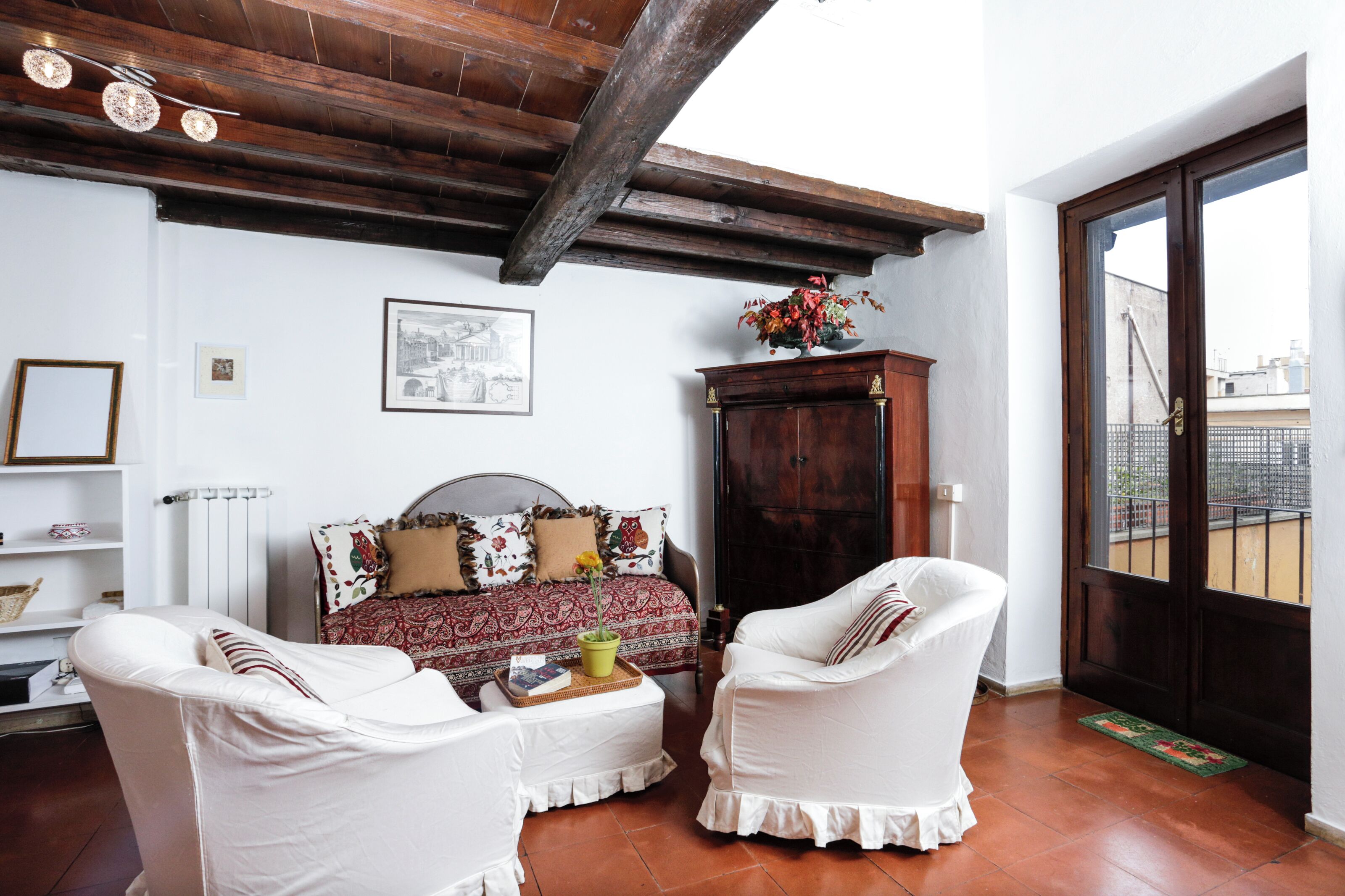 A room with a bed, two white armchairs, a wooden cabinet, and a window with a view of buildings.