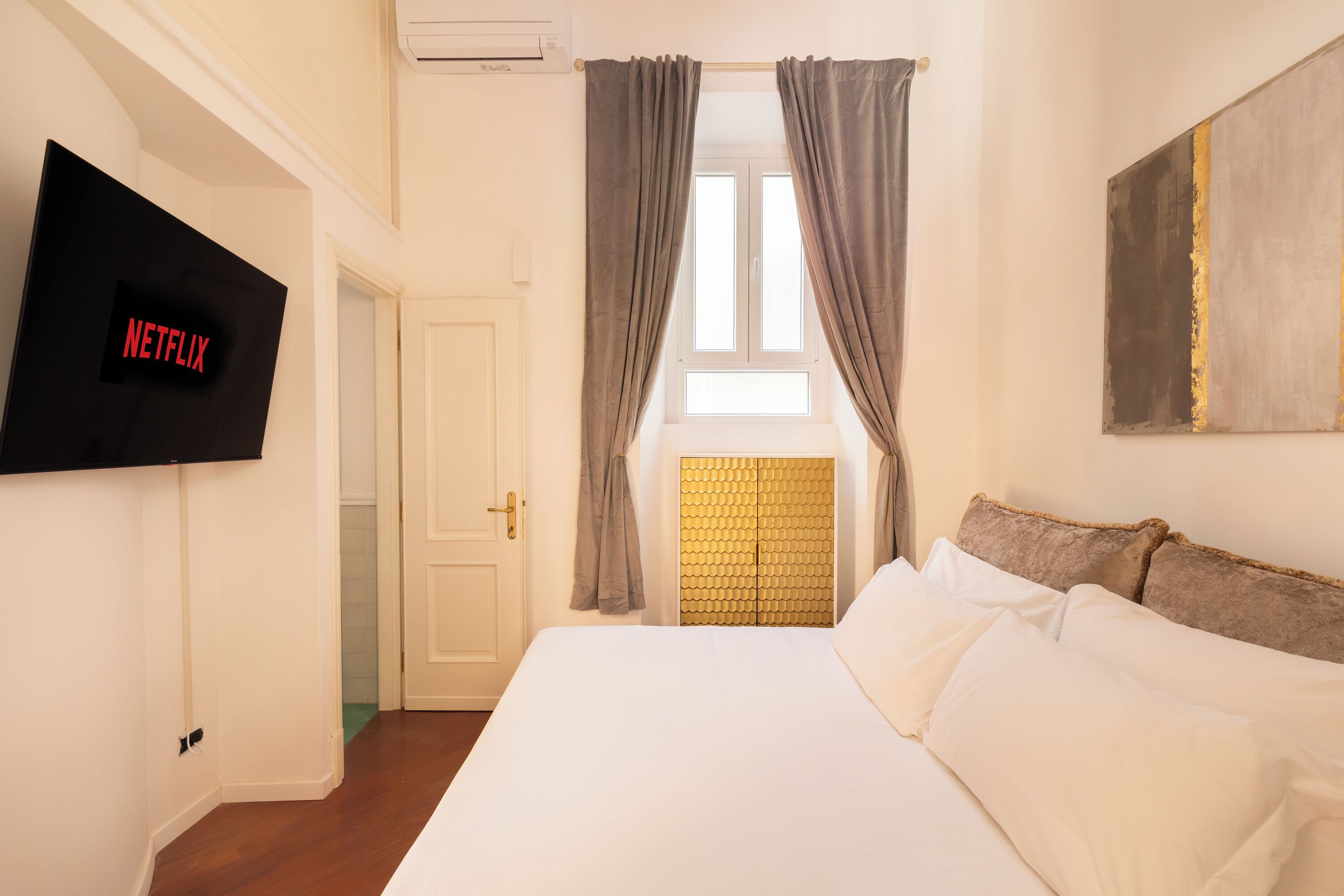 A hotel room with a bed, a TV mounted on the wall, a window with curtains, and a door.