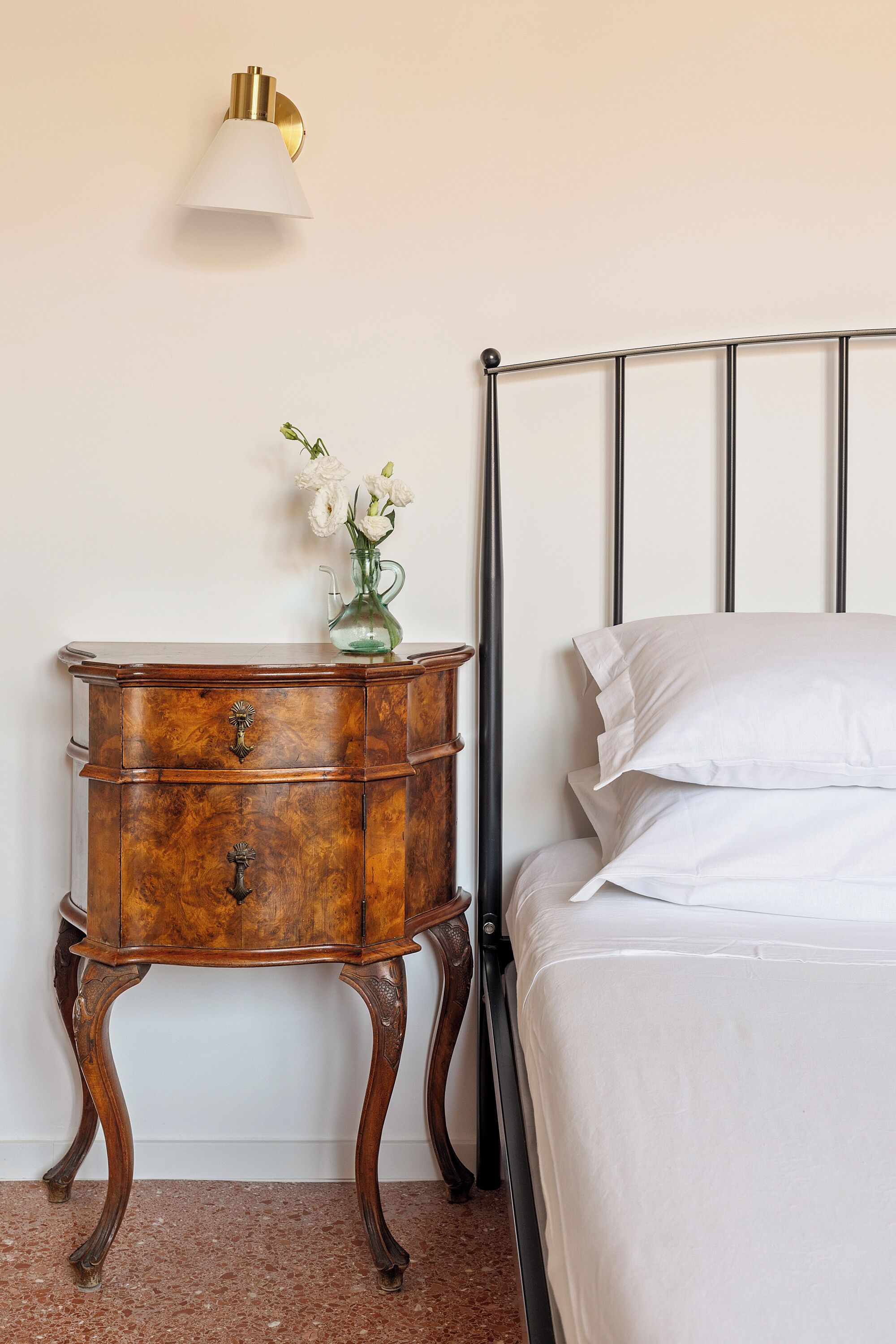 Un dormitorio con una cama, una mesita de noche con un florero y una lámpara de pared.