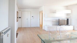 A modern living room with a glass dining table, a flat-screen TV on a white cabinet, and a wooden floor. - Caffarella Park Terraced 2Br Apartment (Rome)