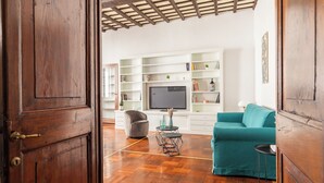 Een woonkamer met een houten vloer, een blauwgroene bank, een grijze fauteuil en een televisie op een witte dressoir.