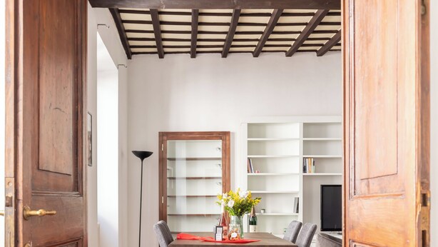 Een eethoek met een houten tafel, stoelen en een bloemenvaas. Achtergrond is een boekenkast en een televisie.